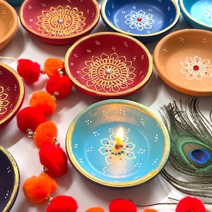Puede incluir: Conjunto de cuencos de cerámica pintados a mano, coloridos, con intrincados diseños florales dorados y blancos. Los cuencos son de varios colores, incluyendo rojo, azul, naranja y turquesa. Una vela encendida está en un cuenco.