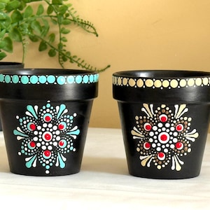 Puede incluir: Dos macetas pintadas de negro con diseños de mandala. Las macetas tienen un borde punteado turquesa y blanco y un diseño de mandala central en rojo, blanco y turquesa. La segunda maceta tiene un borde punteado crema y blanco y un diseño de mandala central en rojo, blanco y marrón.