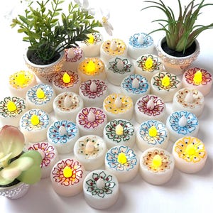 May include: A collection of white LED tea light candles with colorful floral designs. Each candle has a yellow LED light. The candles are arranged with small potted plants, creating a decorative display.