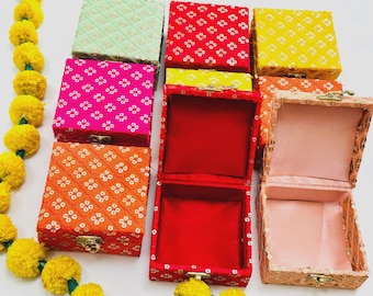 May include: A collection of colorful, square boxes with intricate patterns and gold clasps. The boxes are in various colors, including red, pink, orange, yellow, and green. Some boxes are open, revealing a contrasting interior lining.
