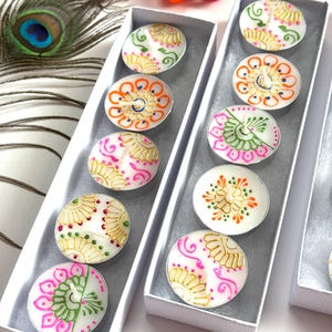 May include: Two white rectangular boxes containing round tea light candles. Each candle features hand-painted designs in gold, pink, orange, and green, including floral and mandala-inspired patterns. The candles are arranged in a single row within each box.