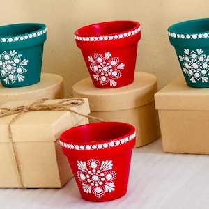 Puede incluir: Maceteros pintados a mano en color verde azulado y rojo con diseños florales y de puntos blancos. Las macetas se exhiben sobre pequeñas cajas redondas de cartón. Una caja de regalo atada con hilo está en primer plano. Estas macetas decorativas son perfectas.