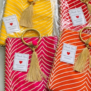 Puede incluir: Cuatro bolsos rectangulares de tela en amarillo, rojo, rosa y naranja, cada uno con un patrón de chevrón dorado, un asa circular dorada, una borla dorada y una etiqueta blanca que dice Thank You for celebrating with us MEERA & RONIT 05/24/2025.