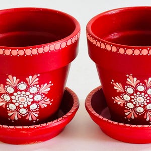 May include: Two matching red terracotta plant pots with attached saucers. Each pot features a hand-painted white and light pink mandala-style floral pattern on the front and a decorative dotted border around the upper rim of the container.