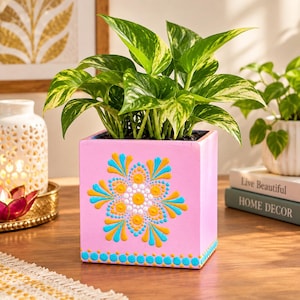 May include: A square pink planter featuring a hand-painted mandala design in yellow, turquoise, and white dots. The planter holds a lush green and yellow variegated pothos plant with heart-shaped leaves, creating a vibrant display.