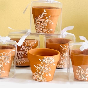 May include: Terracotta-colored flower pots with white floral designs are displayed in clear boxes tied with white ribbons. The pots are small, and the white designs are intricate. The background is a solid yellow.