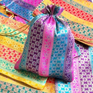 Puede incluir: Una colección de coloridas bolsas con cordón y piezas de tela a juego. Las bolsas y la tela presentan un patrón de rayas con detalles florales en tonos rosa, morado, azul y dorado. Las bolsas se cierran en la parte superior con un cordón dorado.
