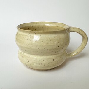May include: A handmade ceramic mug with a distinctive, rounded form and a robust handle. The mug is a light beige colour with small, dark speckles, giving it a rustic look. The mug is designed for holding hot drinks.