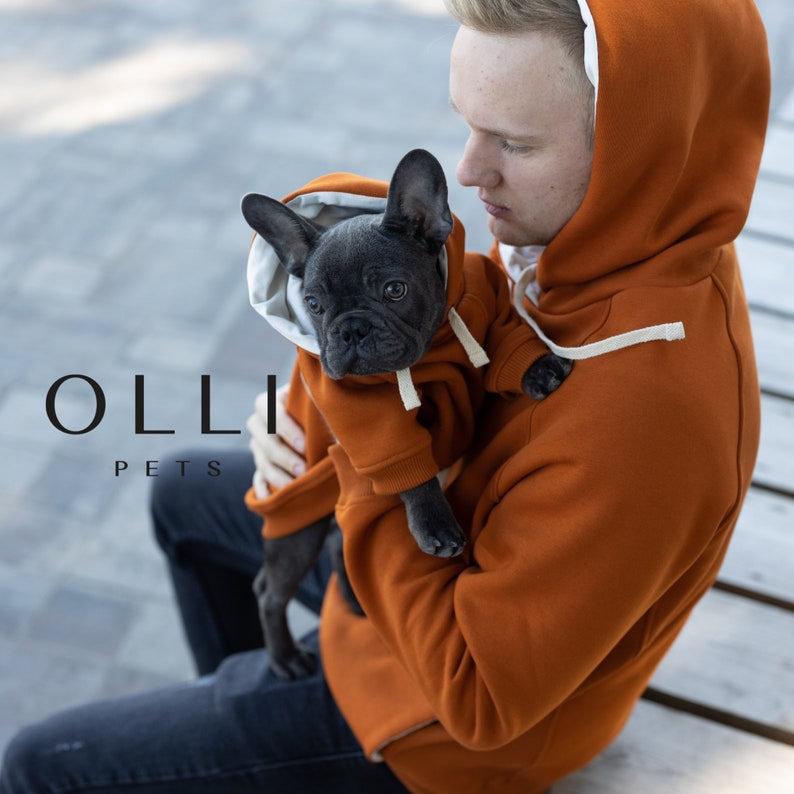 May include: A matching set of burnt orange hoodies with white drawstrings and white interior lining. One hoodie is sized for a person and the other is sized for a small dog. The text OLLI PETS is visible on the left side of the image.