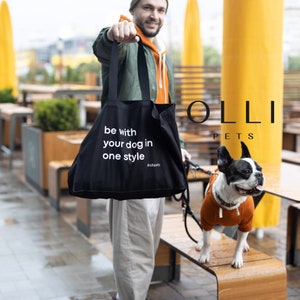 May include: A black tote bag featuring the white printed text "be with your dog in one style #ollipets". The bag has two black shoulder straps and is held in front of a French Bulldog wearing a solid orange sweater with a white drawstring.