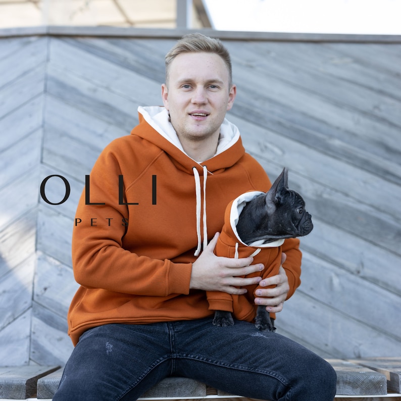 May include: A matching set of burnt orange hoodies with white drawstrings and white inner lining. The larger hoodie features the text OLLI PETS on the chest. A smaller matching hoodie is worn by a black French Bulldog, featuring the same color scheme.