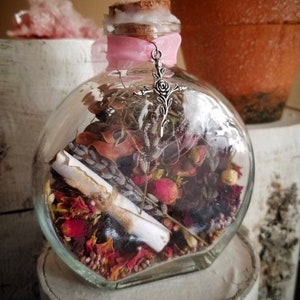 Op de afbeelding: Een heldere glazen fles met een kurkstop en een roze lint om de hals. De fles is gevuld met gedroogde bloemen, kruiden en een opgerold stuk papier. Een zilveren bedel met een kruis en een roosontwerp hangt aan het lint.