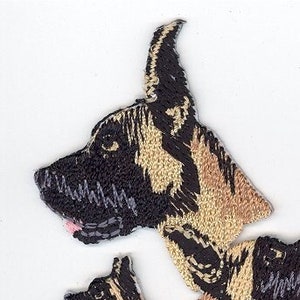 Può includere: Toppa ricamata con un cane marrone e nero. Il cane è in piedi su tutte e quattro le zampe e guarda a destra. La toppa è realizzata in un tessuto morbido e testurizzato.
