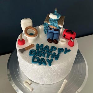 May include: A white cake decorated with fondant figures of a toilet, plunger, toolbox, and a plumber. The cake has blue text that reads "Happy Birthday". The cake is on a silver cake stand.