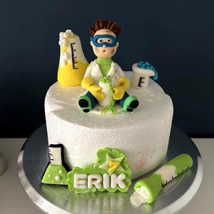 May include: A round white cake decorated with a science theme. It features a fondant figure of a scientist with goggles, lab equipment, and the name "ERIK". The cake has yellow, green, and blue accents.