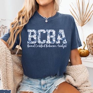 May include: A navy blue t-shirt with a white floral design. The shirt has the text "BCBA Board Certified Behavior Analyst" printed on the front.