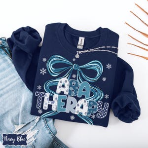 May include: Navy blue sweatshirt with the text "ABA THERAPY" in a decorative font, surrounded by a blue bow and snowflakes. A silver necklace with a pendant is draped on the sweatshirt. The words "Navy Blue" are written on a label.