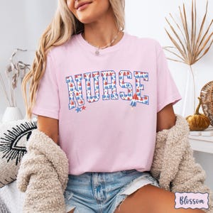 Puede incluir: Camiseta rosa con la palabra "NURSE" en letras blancas con un patr&oacute;n de paletas de hielo rojo, blanco y azul. La camiseta tiene un patr&oacute;n de paletas de hielo rojo, blanco y azul.