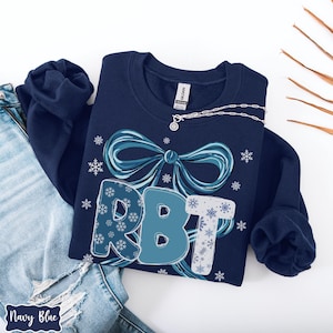 May include: Navy blue sweatshirt with a large bow and the letters "RBT" in shades of blue and white. The letters are decorated with snowflakes. A silver necklace with a pendant is draped on the sweatshirt. The words "Navy Blue" are at the bottom left.