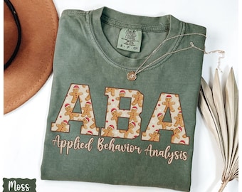 May include: A moss green t-shirt with the letters "ABA" filled with gingerbread men and the words "Applied Behavior Analysis" in a cursive font. A gold necklace and a brown hat are also visible.