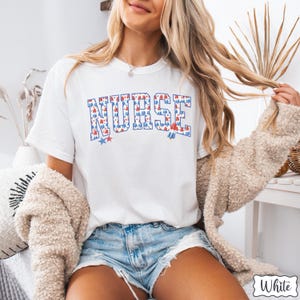 Puede incluir: Camiseta blanca con la palabra "NURSE" impresa en rojo, blanco y azul. Las letras est&aacute;n formadas por un patr&oacute;n de peque&ntilde;as estrellas rojas, blancas y azules.