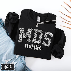 May include: Black sweatshirt with the text "MDS nurse" in a leopard print design. A silver necklace is draped across the front. The sweatshirt is folded on top of a pair of blue jeans.