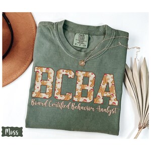 May include: A moss green t-shirt with the letters "BCBA" in a gingerbread man pattern. The text "Board Certified Behavior Analyst" is below the letters. A gold necklace and a brown hat are next to the shirt.