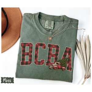 May include: A moss green t-shirt with the letters "BCBA" in a red and black plaid pattern. A red truck carrying a Christmas tree is printed below the letters. A gold necklace and a brown hat are also visible.
