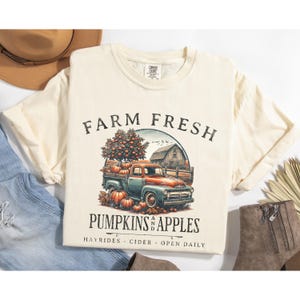 May include: A cream colored t-shirt with a vintage graphic of a red and blue pickup truck carrying pumpkins and apples. The text "FARM FRESH PUMPKINS APPLES HAYRIDES - CIDER - OPEN DAILY" is printed on the shirt.