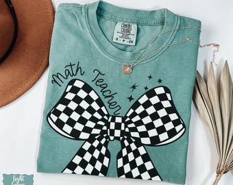 Può includere: T-shirt verde chiaro con la scritta "Math Teacher" e un grande disegno a fiocco a scacchi bianchi e neri. Una collana d'argento con un piccolo ciondolo è drappeggiata sulla maglietta. La maglietta è piegata su una superficie bianca.