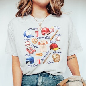May include: A white t-shirt with a baseball-themed design. The design features a variety of baseball-related items, including a baseball bat, a baseball glove, a baseball cap, a baseball, a hot dog, a pretzel, a soda can, and a popcorn box. The words "At Bat", "Walk", "Strike", "Home Run", "Single", "Dugout", "Triple", "Foul Ball", "Strike Out", "Inning", "Base", and "Double" are written around the design.
