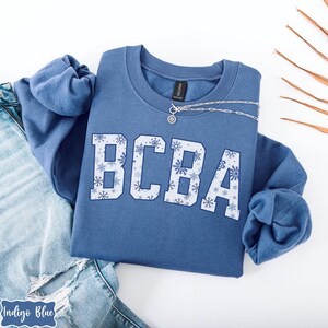 May include: A blue crewneck sweatshirt with the letters "BCBA" in a white, snowflake-filled design. A silver necklace with a pendant is draped on the sweatshirt. The sweatshirt is folded on top of a pair of blue jeans.