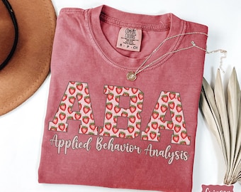 May include: A crimson red t-shirt with the letters "ABA" in white outlined in black. The letters are filled with a pattern of red strawberries. Below the letters is the text "Applied Behavior Analysis" in white outlined in black.