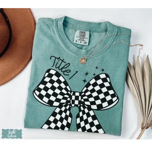 May include: Light green t-shirt with a large black and white checkered bow graphic. The text "Title 1" is above the bow. A gold necklace and a brown hat are also visible.