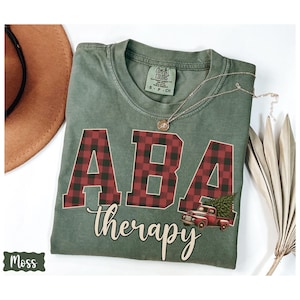 May include: Moss green t-shirt with the words "ABA therapy" in a script font. The letters "ABA" are in a red and black plaid pattern. A red truck carrying a Christmas tree is on the right side of the shirt. A gold necklace is on the shirt.