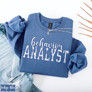 May include: A blue sweatshirt with the words "behavior ANALYST" in white script and block letters. The word "behavior" is in script, and "ANALYST" is in block letters with snowflake accents. A silver necklace with a pendant is draped on the sweatshirt.