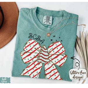 May include: A light green Comfort Colors t-shirt with a Christmas bow design. The bow is white with red stripes and green sprinkles, and a Christmas tree in the center. The text "BCBA" is above the bow. A gold necklace is draped across the shirt.