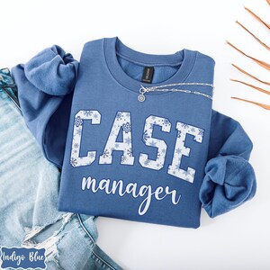 May include: A blue sweatshirt with the words "CASE manager" in white block letters with snowflake patterns. A silver necklace with a pendant is draped on the sweatshirt. The sweatshirt is folded on top of a pair of blue jeans.