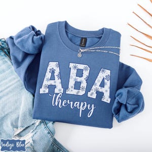May include: A blue sweatshirt with the text "ABA therapy" in white script. The letters "ABA" are filled with a snowflake pattern. A silver necklace with a pendant rests on the sweatshirt. The sweatshirt is folded on top of a pair of blue jeans.