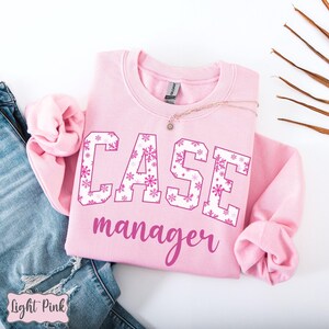 May include: A light pink sweatshirt with the words "CASE manager" in a script font. The letters "CASE" are white with a pink snowflake pattern. A gold necklace is draped across the sweatshirt. The words "Light Pink" are in the bottom left corner.