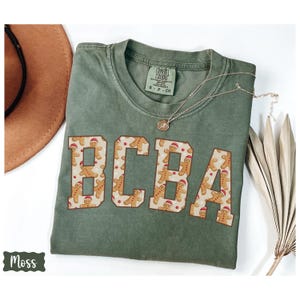 May include: A moss green t-shirt with the letters "BCBA" in a gingerbread man pattern. The shirt has a crew neck and short sleeves. A gold necklace with a pendant is draped on the shirt. A brown hat is visible in the background.