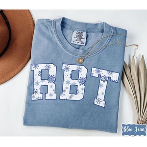 May include: A blue Comfort Colors t-shirt with the letters "RBT" in white, filled with blue snowflakes. A gold necklace and a brown hat are visible. The words "Blue Jean" are in a blue script font on the bottom right.