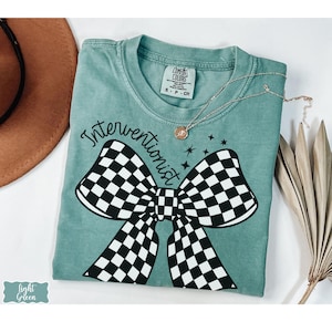 May include: Light green t-shirt with the word "Interventionist" in black script above a large black and white checkered bow. A delicate gold necklace with a pendant rests on the shirt. The shirt has a Comfort Colors tag.