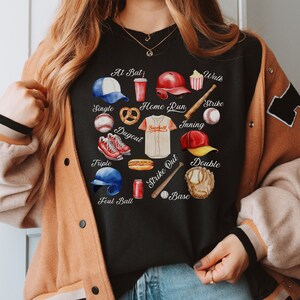 May include: Black t-shirt with a heart-shaped design featuring baseball-themed illustrations and text. The text includes "At Bat", "Walk", "Single", "Home Run", "Strike", "Inning", "Dugout", "Triple", "Foul Ball", "Strike Out", "Double", and "Base".