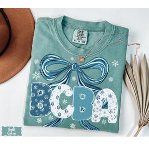 May include: Light green t-shirt with a large blue bow design and the letters "BCBA" in a snowflake pattern. A delicate necklace and a brown hat are also visible. The shirt is folded neatly on a white surface.