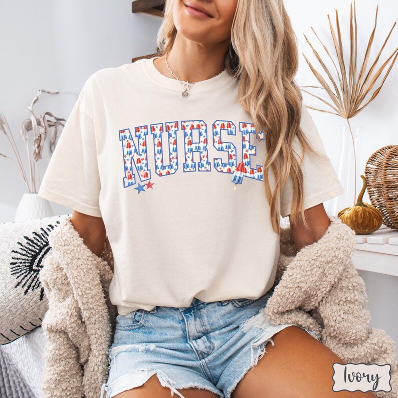Puede incluir: Una camiseta blanca con la palabra "NURSE" impresa en rojo, blanco y azul. Las letras est&aacute;n formadas por un patr&oacute;n de peque&ntilde;as estrellas rojas, blancas y azules.