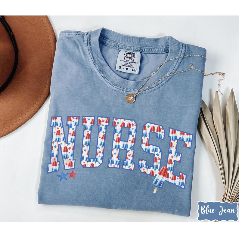 Puede incluir: Una camiseta azul claro con la palabra "NURSE" impresa en letras blancas grandes. Las letras est&aacute;n decoradas con un patr&oacute;n de paletas de hielo rojas, blancas y azules y estrellas. La camiseta est&aacute; hecha por Comfort Colors.