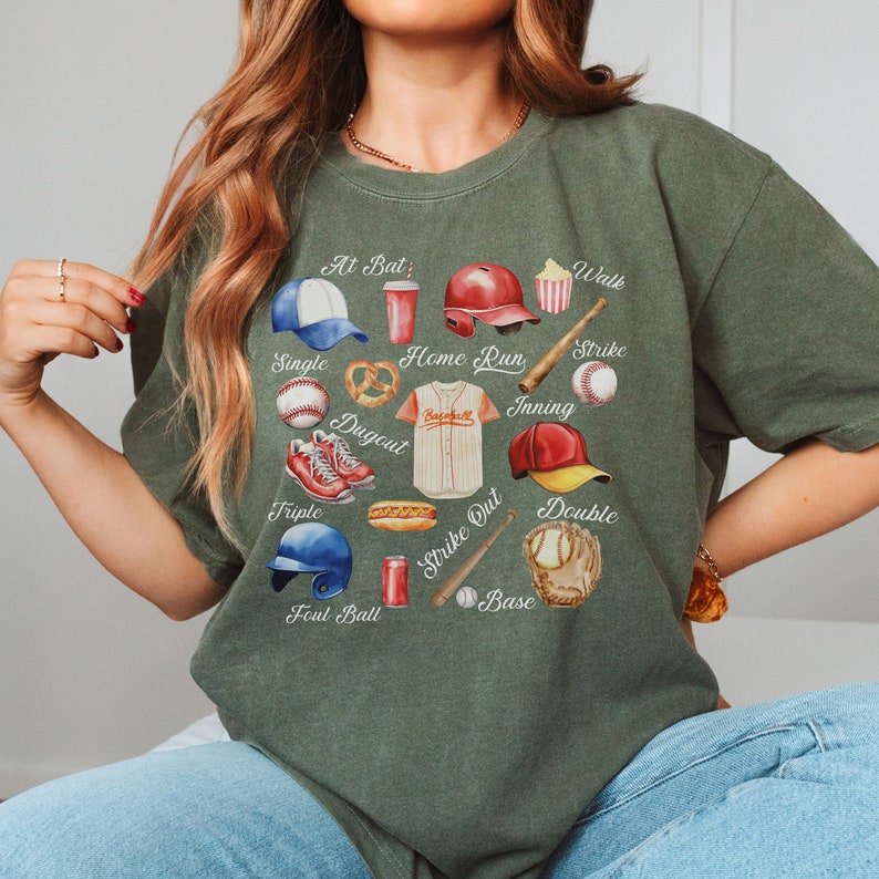 May include: A green t-shirt with a baseball-themed design. The design features various baseball-related items, including a baseball glove, a bat, a baseball, a baseball cap, a hot dog, a pretzel, a can of soda, and a baseball uniform. The words "At Bat", "Walk", "Strike", "Single", "Home Run", "Inning", "Dugout", "Triple", "Foul Ball", "Strike Out", "Double", and "Base" are written around the design.
