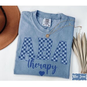 May include: A blue Comfort Colors t-shirt with the words "ABA therapy" in a dark blue font. The letters "ABA" are in a checkered pattern. A gold necklace and a brown hat are visible.