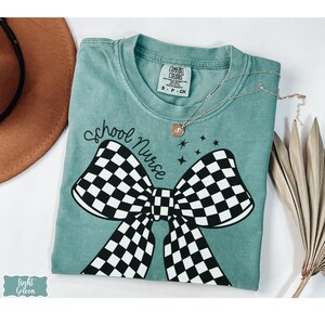 May include: A light green t-shirt with the words "school nurse" above a large black and white checkered bow. A gold necklace with a pendant is draped across the shirt. The shirt is labeled "Comfort Colors" and is size S-P-CH.